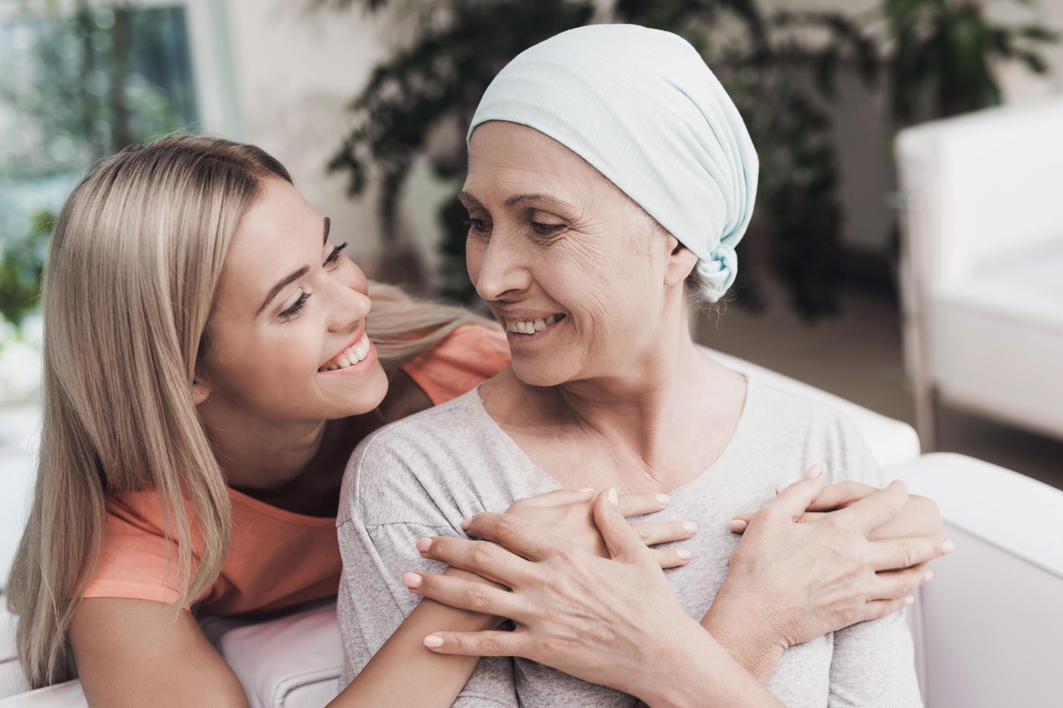 A woman with cancer is sitting on a white sofa next to her daughter. A girl is hugging a woman. They are sitting in the lobby of a modern clinic. Around them flowerpots with flowers.